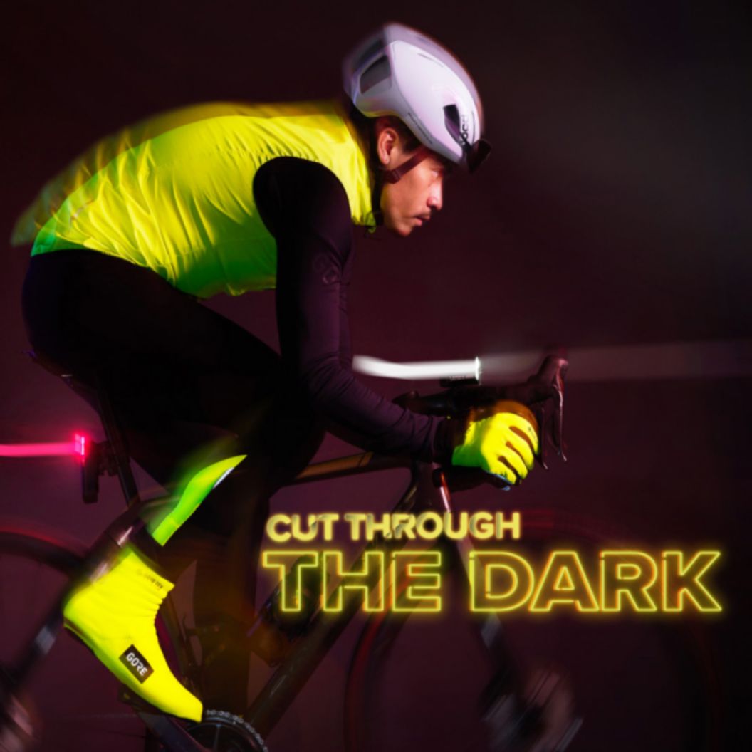 A glowy and slightly motion-blurred image of a road rider in heavy winter apparel riding with lights in a pitch-black scene. Their high-vis and reflective kit catches the light. “Cut through the dark” text in neon lights is shown. 